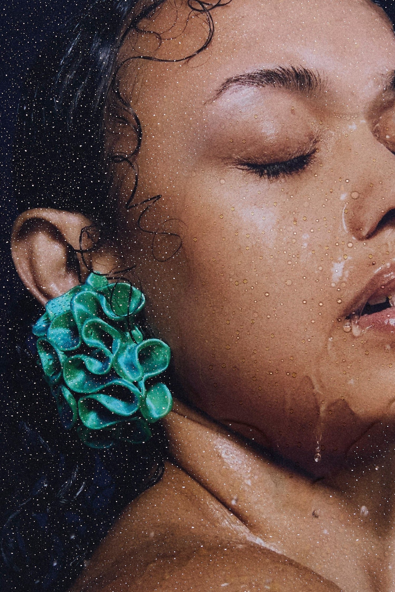 Close-up of a model's face with water droplets, wearing Bhumi's 'Bathe in the Waters' collection. The model showcases the Marina earring and earcuff in Satin Blue/Green Multitone, reflecting the coral-inspired, 3D-printed design and bold aesthetic of the sustainable luxury jewelry.