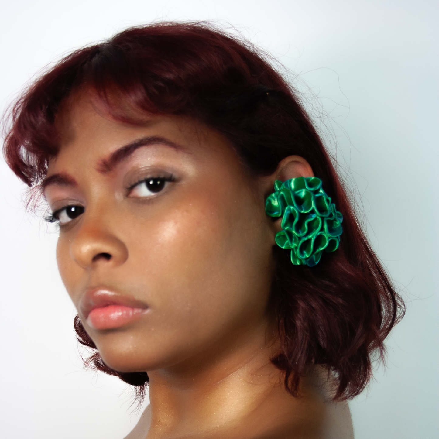 A headshot of a muse showcasing the Marina earcuff in satin blue/green multitone from Bhumi's 'Bathe in the Waters' collection. The coral-inspired, 3D-printed design stands out with its textured, oceanic hues.