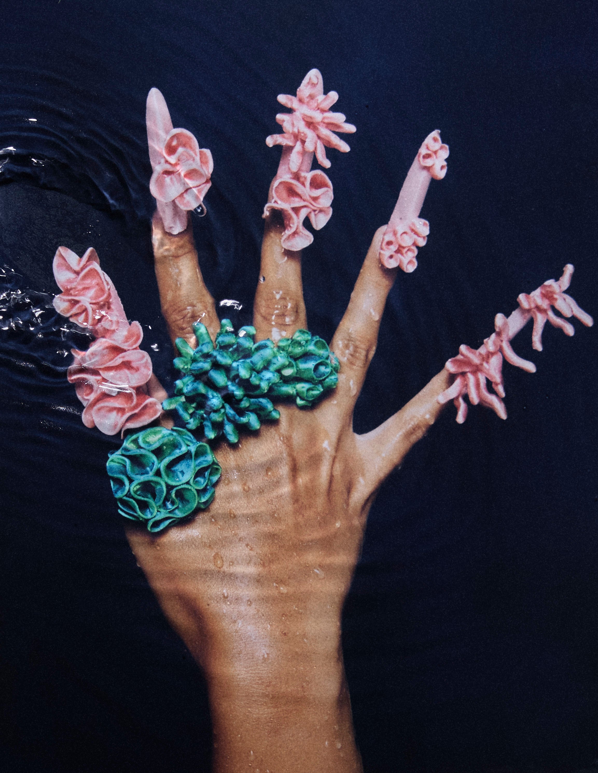 Close-up of a hand with satin pink nails, wearing coral-inspired 3D-printed rings from Bhumi's 'Bathe in the Waters' collection. The rings feature a Satin Blue/Green Multitone design, showcasing the intricate craftsmanship of the sustainable luxury jewelry.