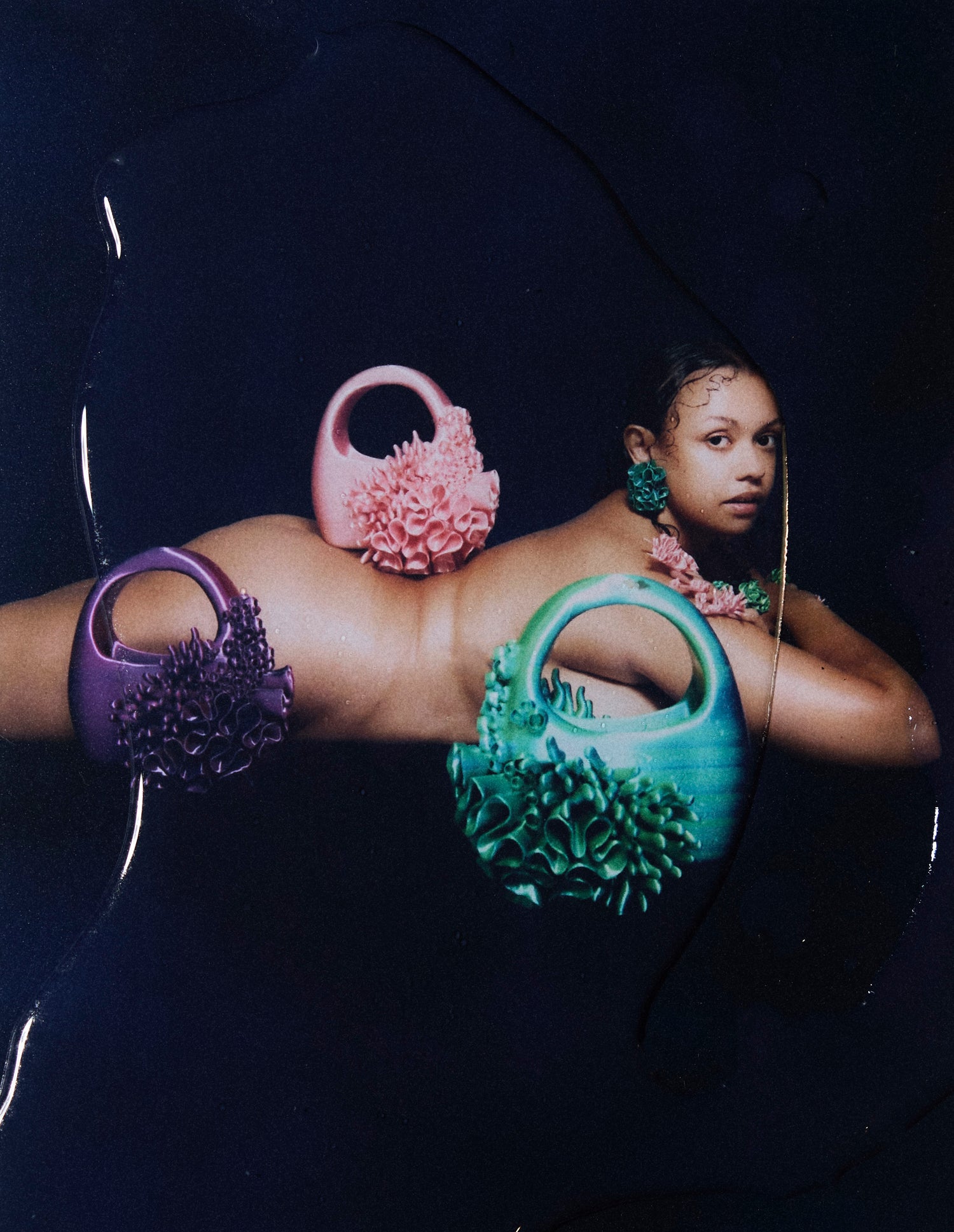 Naked model lying on her front, head turned to look into the camera, wearing Bhumi's 'Bathe in the Waters' collection. The model showcases coral-inspired 3D-printed accessories, including the Siren bag in Satin Pink, Satin Blue/Green Multitone, and Satin Purple/Red, along with Marina earrings and rings. Her pink nails complement the intricate, sustainable luxury jewelry, capturing the essence of the collection.