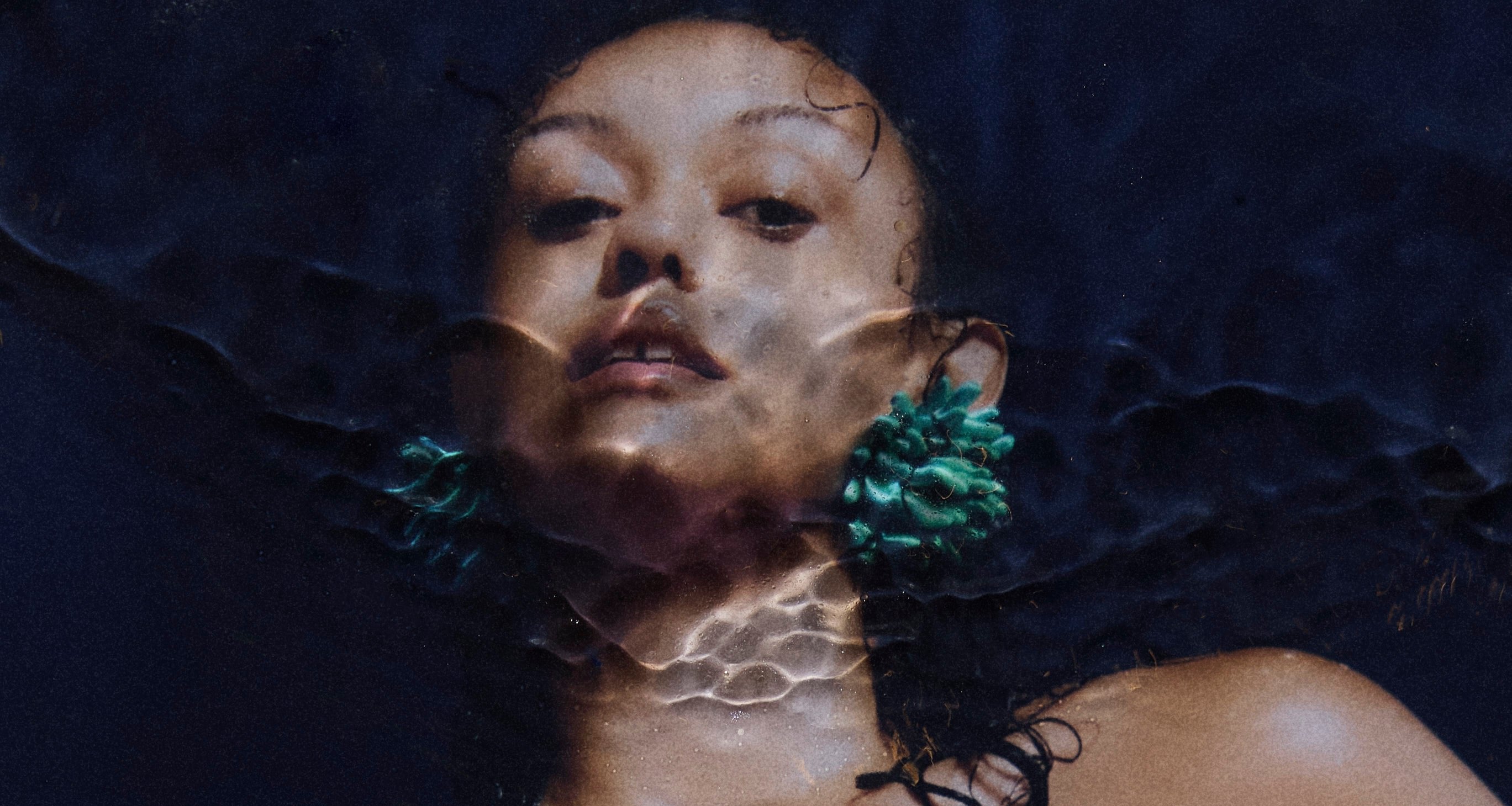 Model wearing Bhumi Clothing's 'Bathe in the Waters' collection, featuring coral-inspired 3D-printed Spheara and Marina rings in Satin Blue/Green Multitone, Anemone earrings, and an ear cuff. The model, with satin pink nails, holds her chest in a front-facing pose, showcasing the intricate design of the sustainable luxury jewelry.